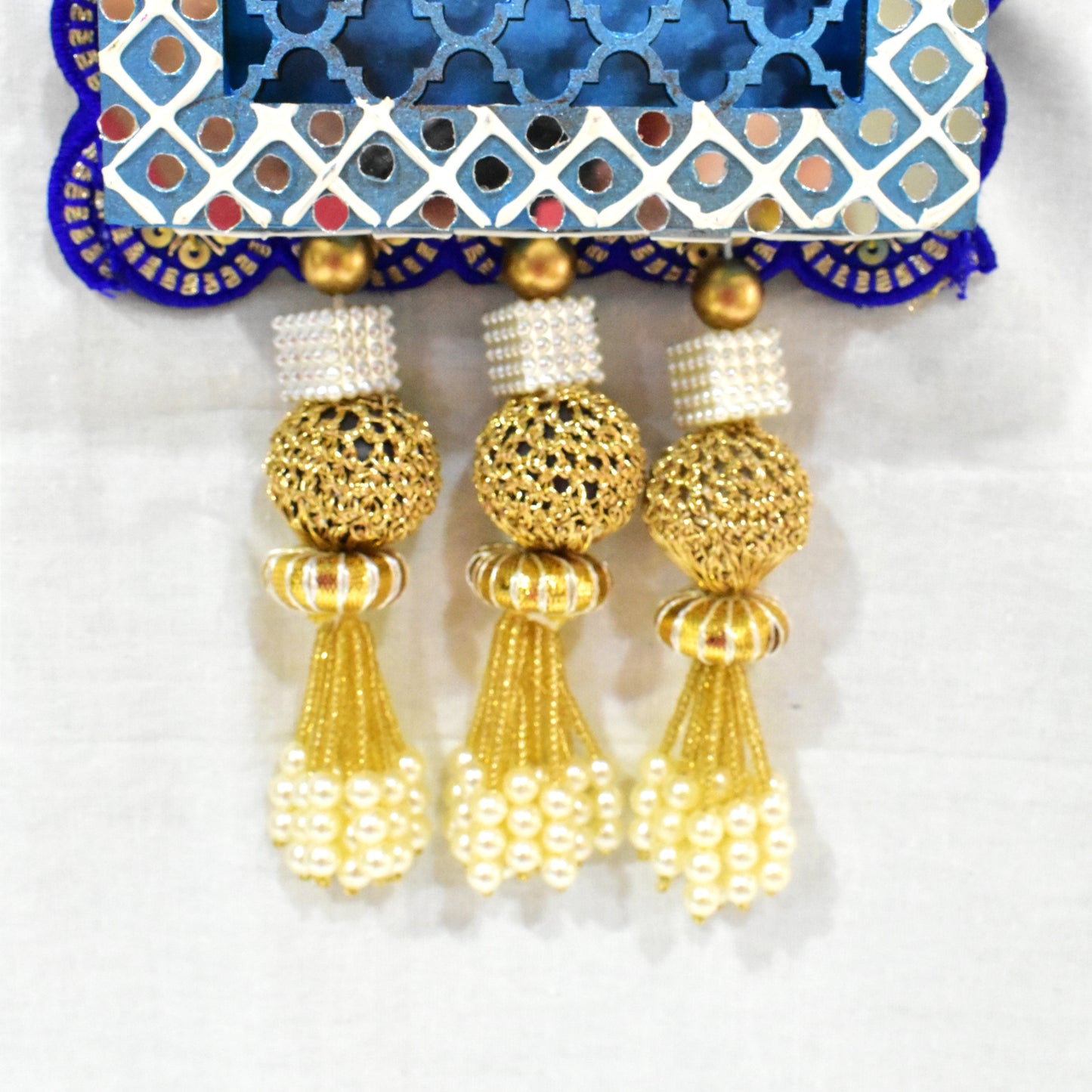 A Handcrafted Kamdhenu Jharokha Wall Hanging with a blue and white geometric patterned frame, featuring a red and gold cow figurine, and pearl-like decorations at the bottom.