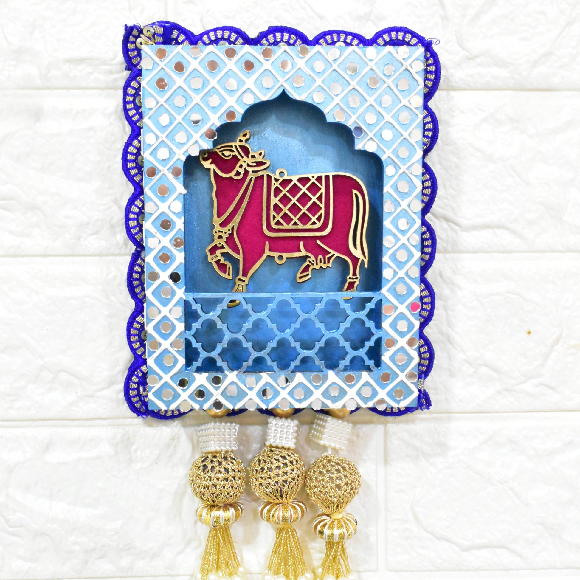 Jharokha Wall Hanging with a blue and white geometric patterned frame, featuring a pink and gold cow figurine, and pearl tassels.
