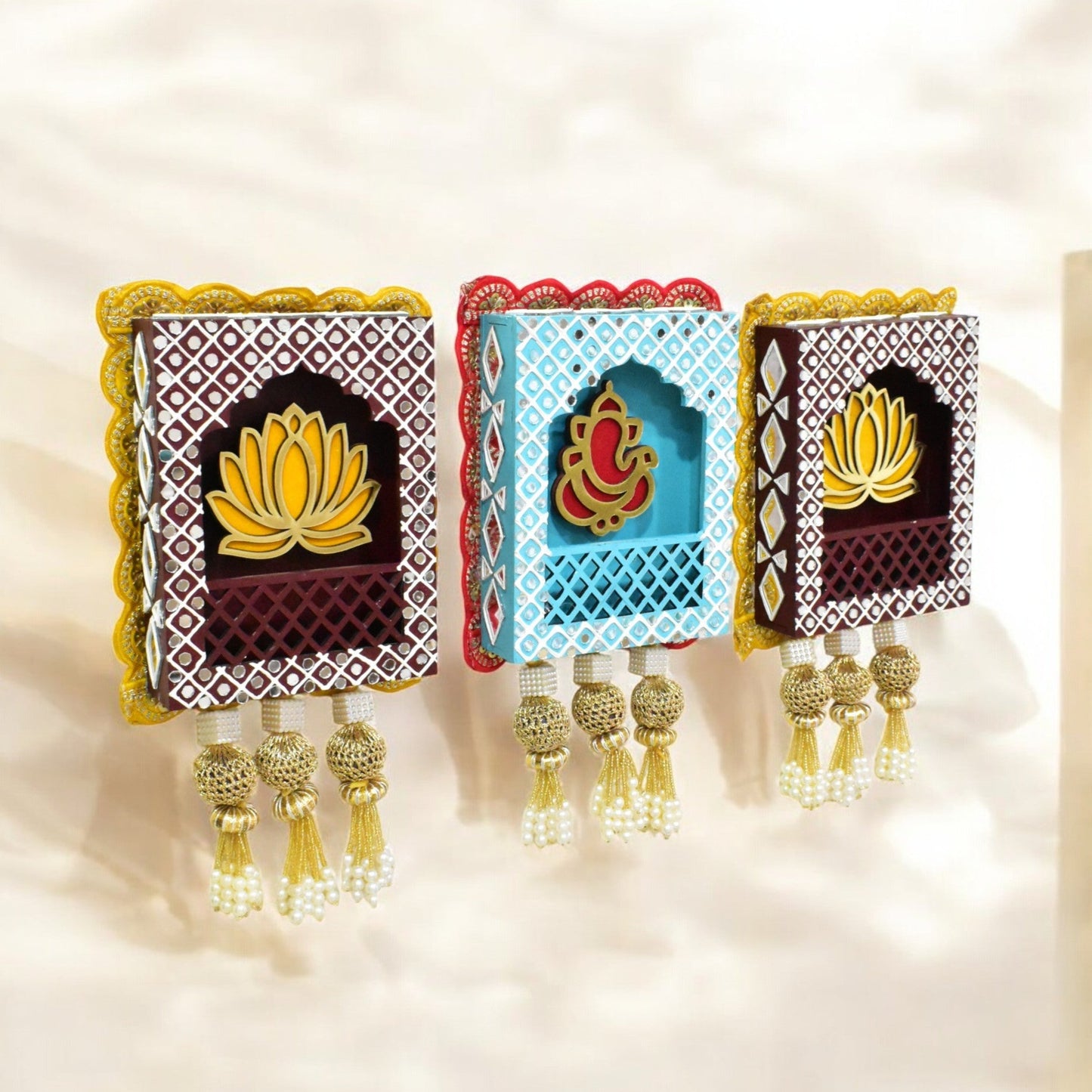 A set of three wall hangings featuring Lord Ganesha with a lotus flower design, in blue, brown, and gold colors, with pearl tassels.