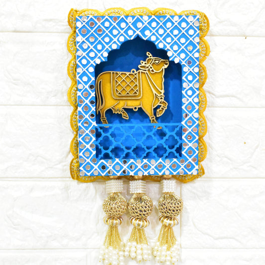A Handcrafted Kamdhenu Jharokha Wall Hanging featuring a golden cow within a blue and yellow detailed frame, with tassels at the bottom.