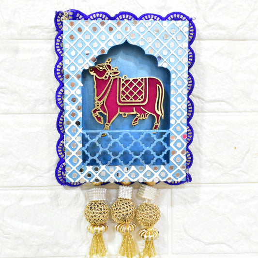 A Handcrafted Kamdhenu Jharokha Wall Hanging with a blue and white geometric patterned frame, featuring a red and gold cow figurine, and pearl-like decorations at the bottom.