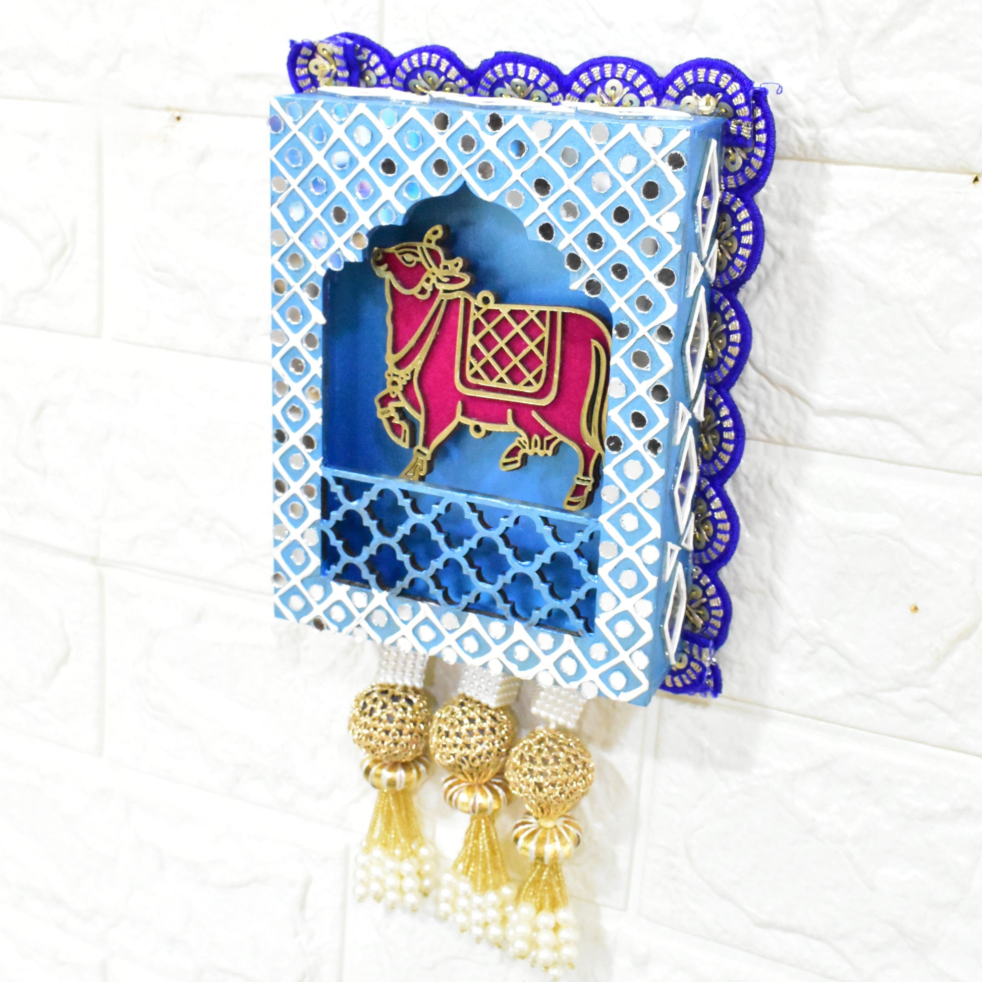 A Handcrafted Kamdhenu Jharokha Wall Hanging with a blue and white geometric patterned frame, featuring a red and gold cow figurine, and pearl-like decorations at the bottom.