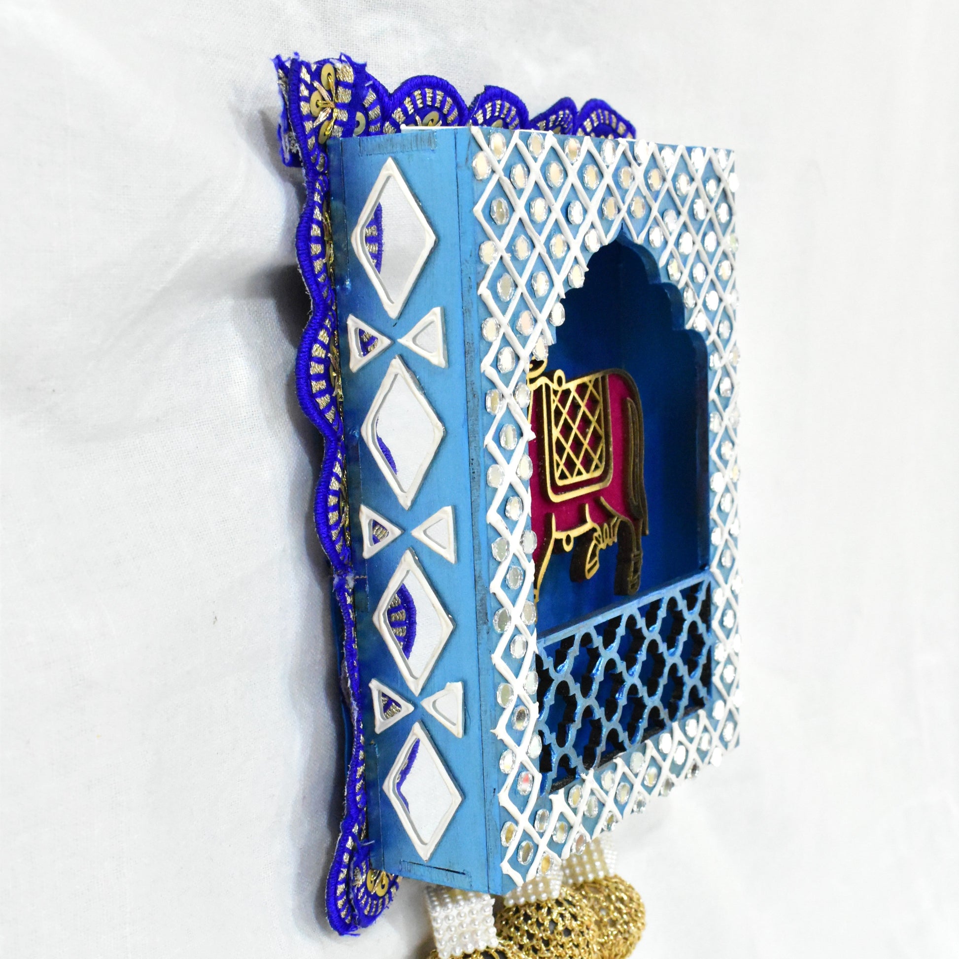 A Handcrafted Kamdhenu Jharokha Wall Hanging with a blue and white geometric patterned frame, featuring a red and gold cow figurine, and pearl-like decorations at the bottom.