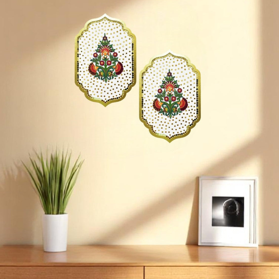 Two decorative wall art pieces on a beige wall above a wooden cabinet with a plant and framed picture.