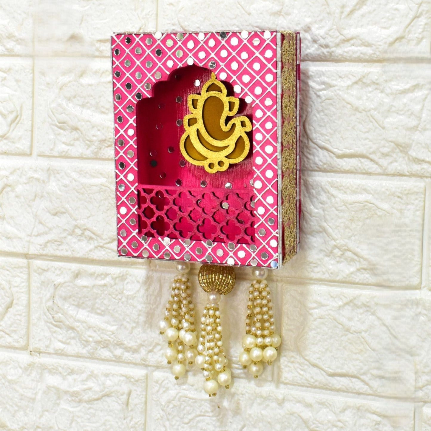 Decorative wall hanging jharokha in pink, with gold Ganesha in the middle, pearl tassels and mirrors on a white textured wall.