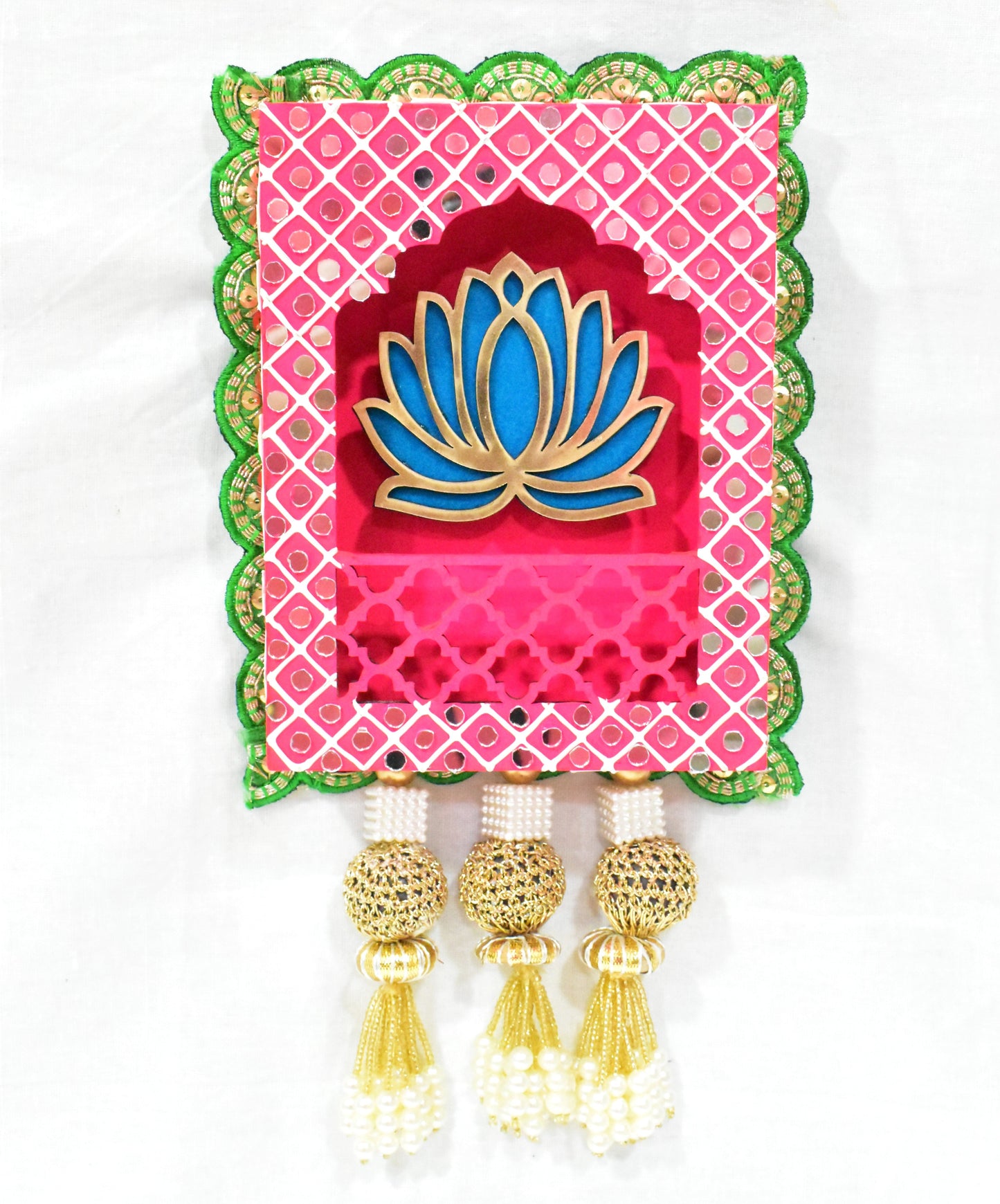 A Lotus Jharokha Wall Hanging with a blue lotus flower on a pink and green patterned background, with gold-colored decorative elements and three gold & pearl tassels .