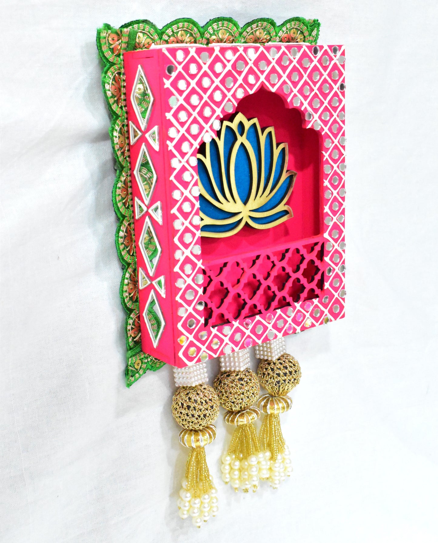 A Lotus Jharokha Wall Hanging with a blue lotus flower on a pink and green patterned background, with gold-colored decorative elements and three gold tassels at the bottom.