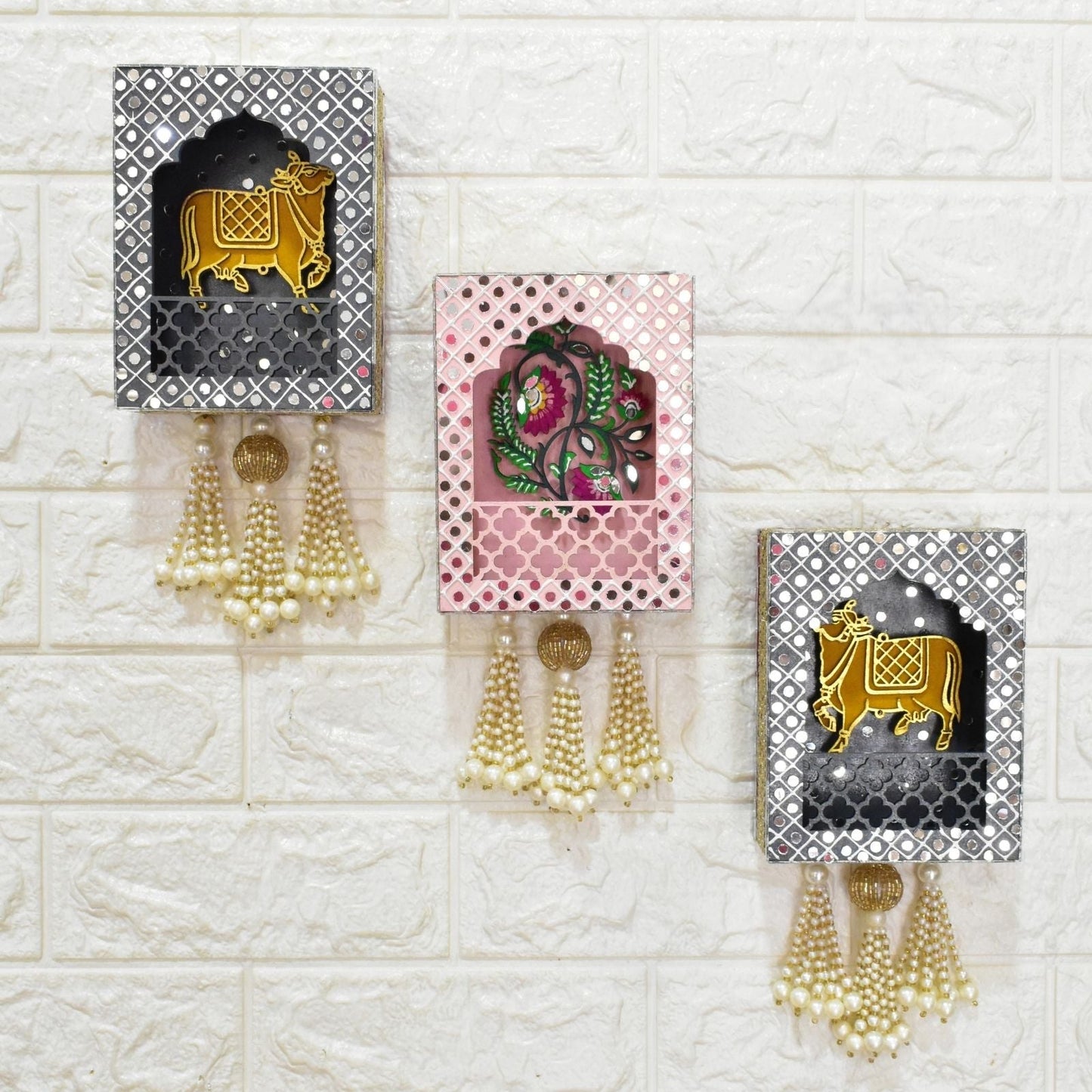 Decorative wall hanging jharokhas with intricate designs and tassels on a textured white wall.