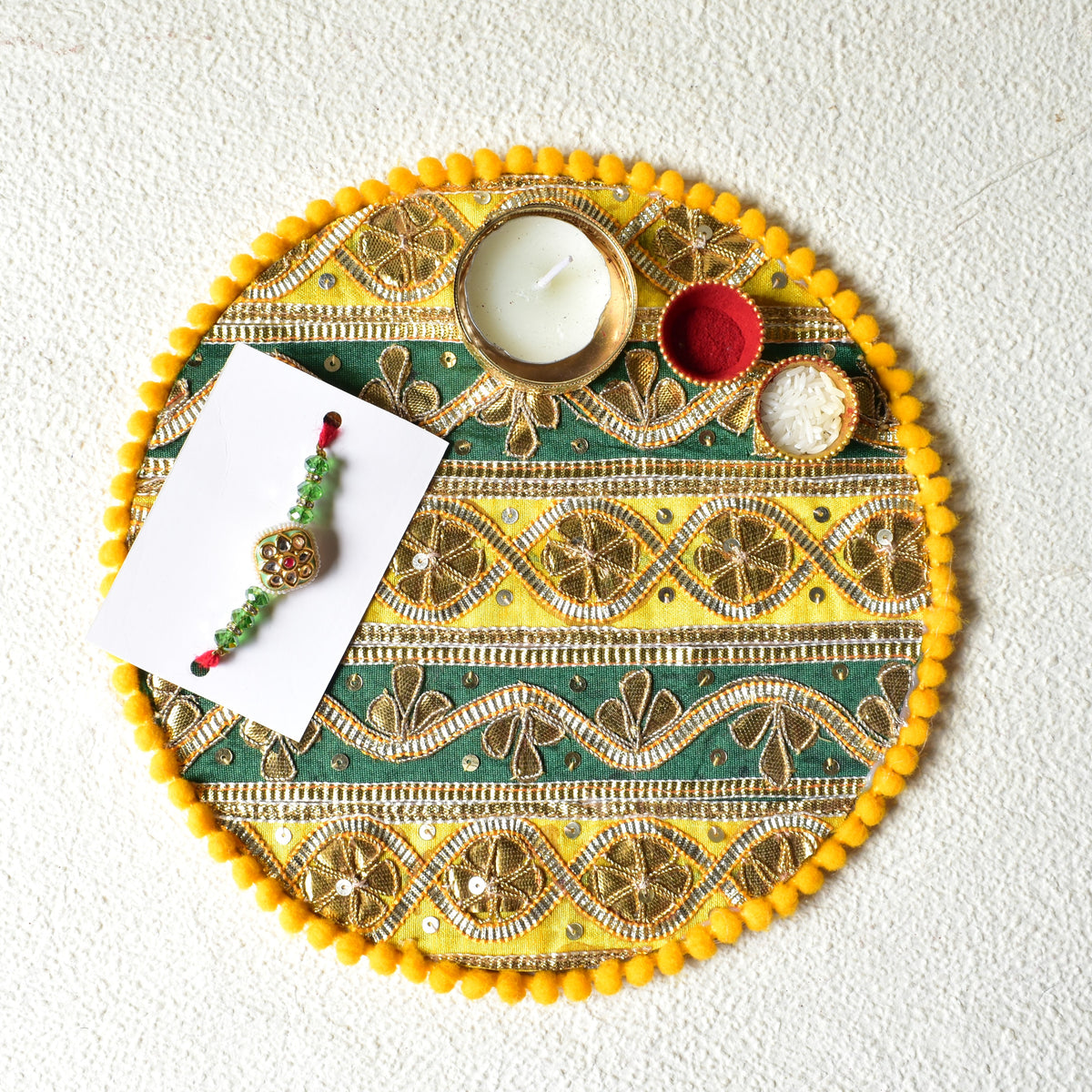 A circular decorative tray with yellow pom-pom trim and green-and-golden yellow embroidered patterns, styled with a small Rakhi on a white card, a candle holder, and two small bowls on a textured off-white background.